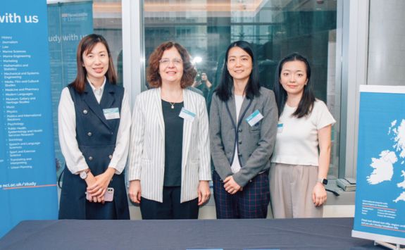 Cristina Neesham standing with three Newcastle University Business School alumni at a formal event. They are positioned in front of a backdrop with educational information and a map of the UK,